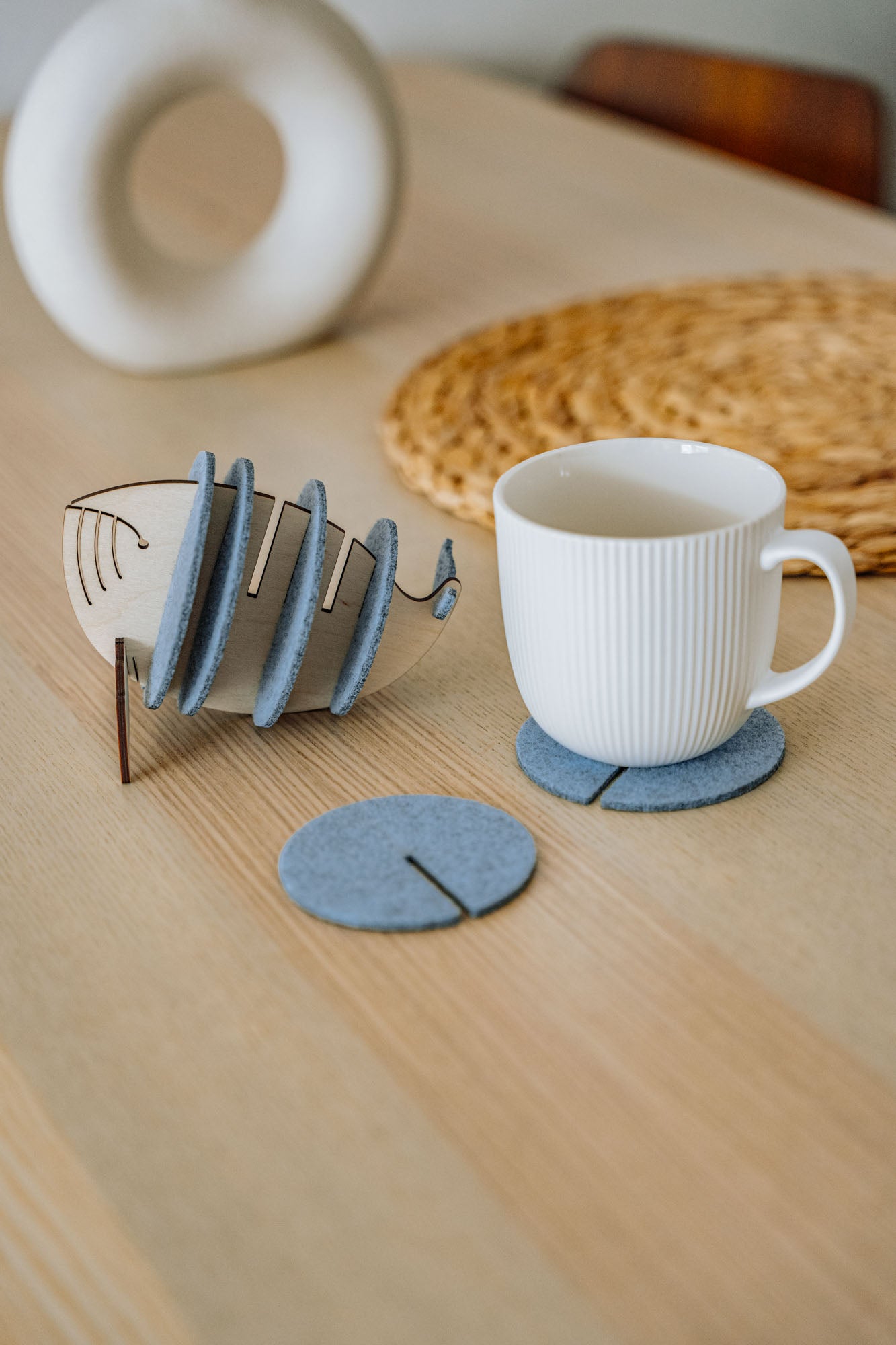 A white ribbed mug sits on a round gray felt coaster, designed to protect surfaces. Nearby, more Whale Coasters are stacked in a fish-shaped holder, with woven and ceramic decor adding home charm in the background.