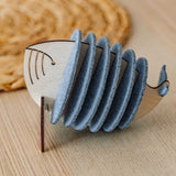 A wooden Whale Coasters holder shaped like a whale, with several round grey coasters inside, sits on a light wooden table near a woven mat—perfect to protect surfaces and add home charm.