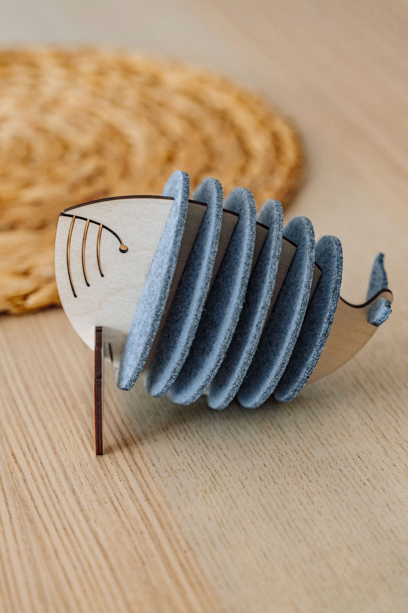 A wooden Whale Coasters holder shaped like a whale, with several round grey coasters inside, sits on a light wooden table near a woven mat—perfect to protect surfaces and add home charm.
