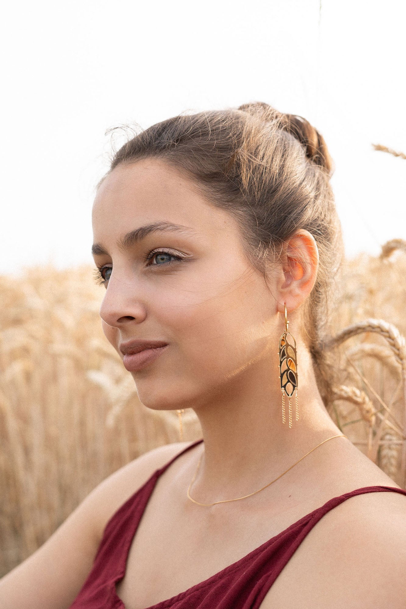A woman with light brown hair in a bun, wearing a sleeveless burgundy top, gold necklace, and Wheat Statement Earrings, sits in a wheat field, looking thoughtfully to the side—her standout style capturing quiet elegance.