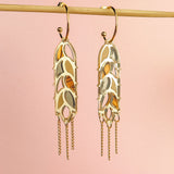 A pair of Wheat Statement Earrings with cut-out leaf patterns and dangling chain accents hang from a horizontal rod against a soft pink background, showcasing standout style.