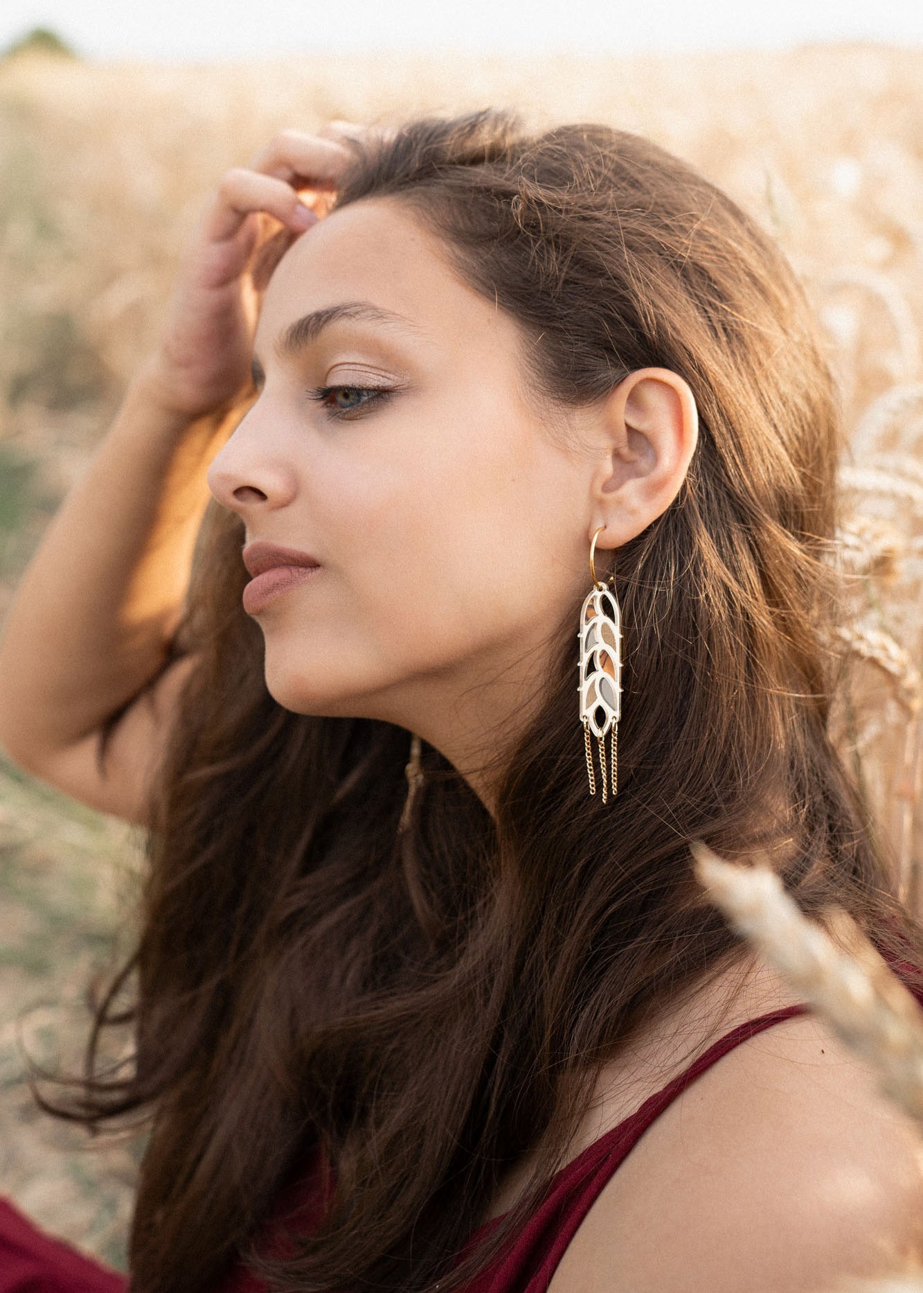 A young woman with long brown hair sits in a wheat field, wearing a burgundy dress and Wheat Statement Earrings. She gazes thoughtfully to the side, lightly touching her hair, her standout style enhanced by the bold design of her earrings.