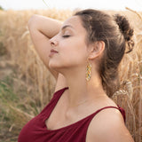 A young woman in a burgundy dress sits in a wheat field with her eyes closed, one arm raised behind her head. She wears Wheat Statement Earrings and a delicate necklace, appearing relaxed and peaceful in the natural setting with standout style.